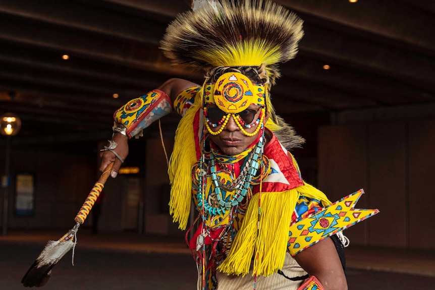 A man poses in bent over stance in sunglasses and bright yellow, brown, red and turquoise Native American regalia.