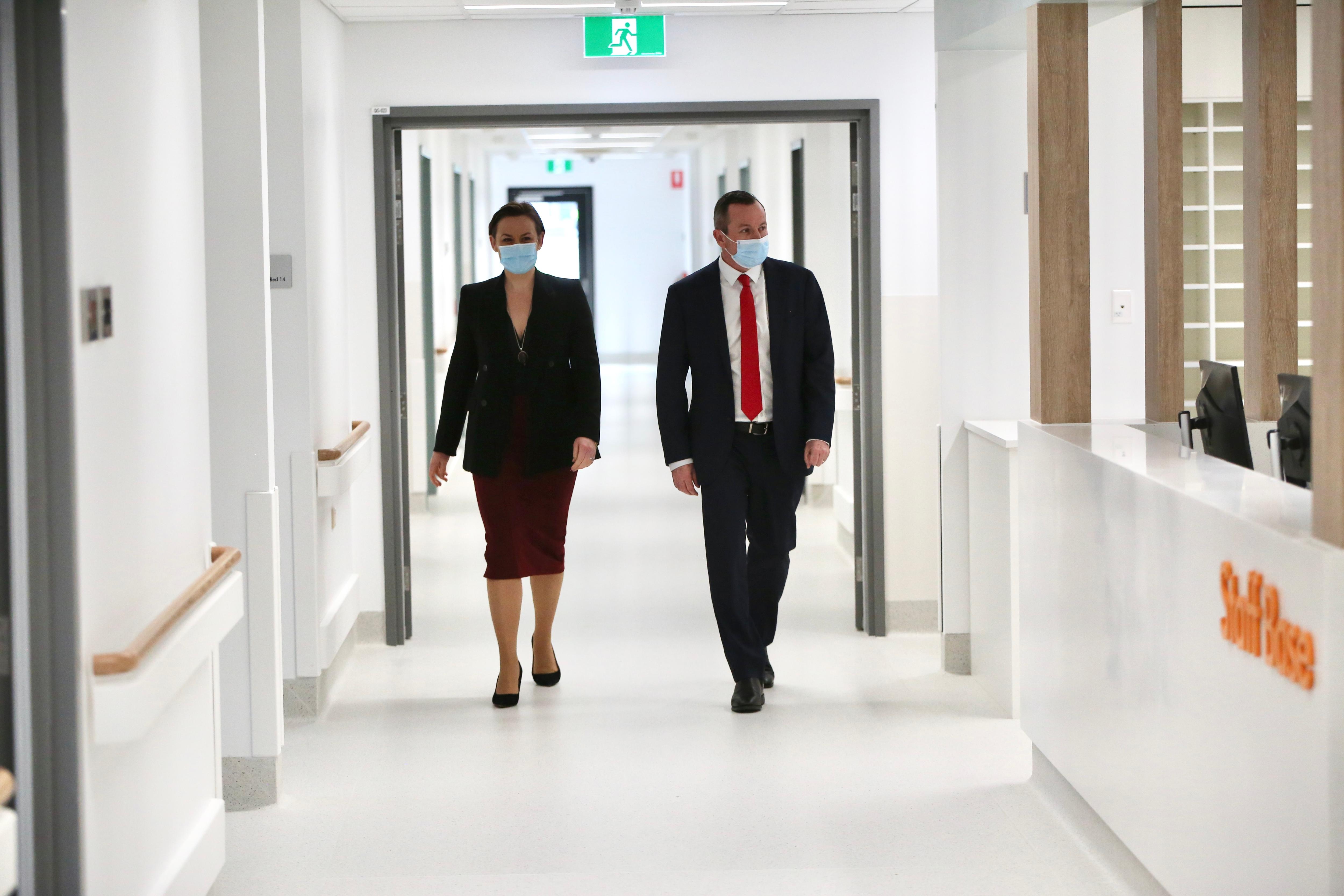 A well dressed man and woman wearing face masks walk down a hospital corridor. 