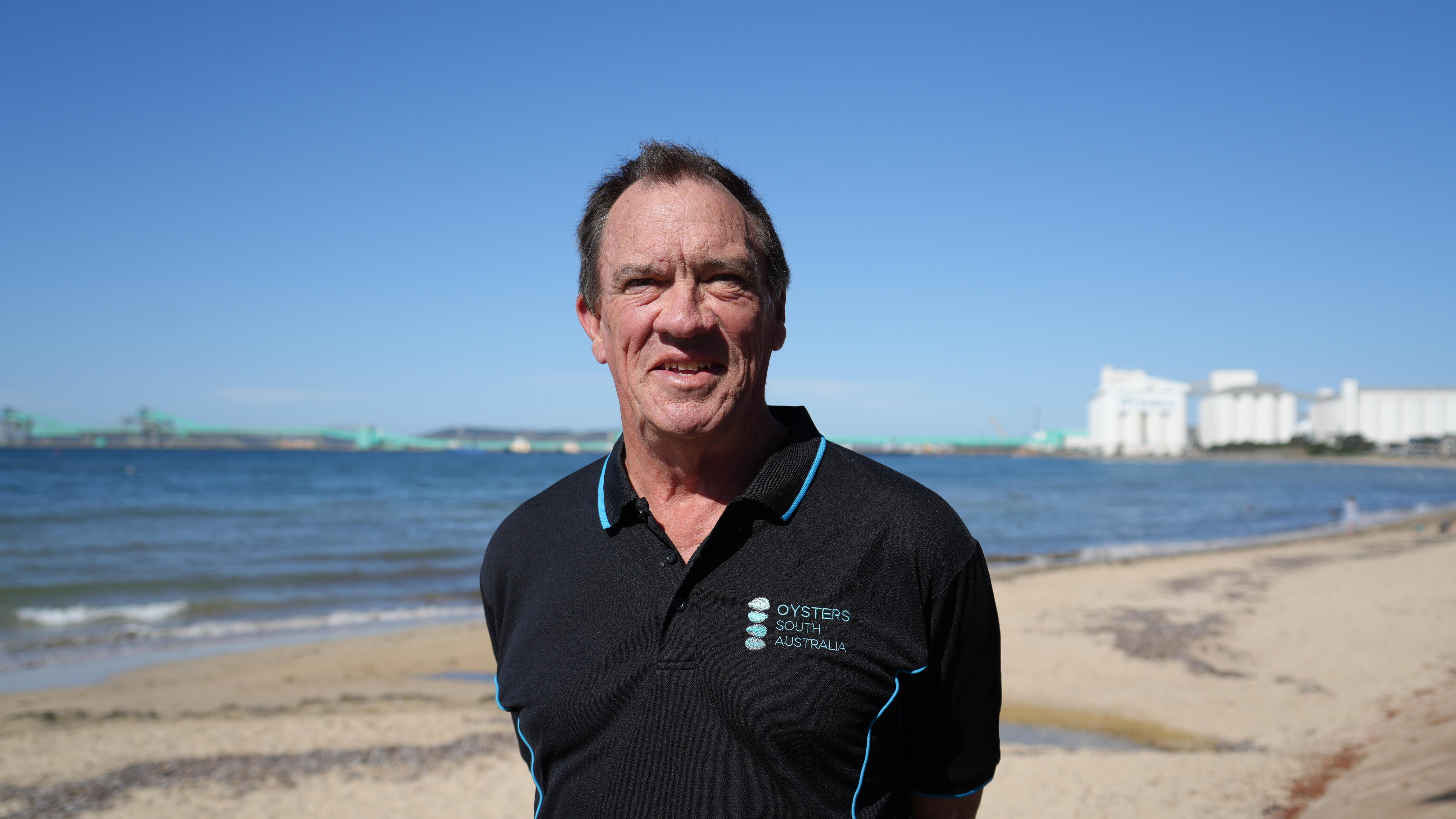 A man smiling at the camera, with a beach landscape behind him.