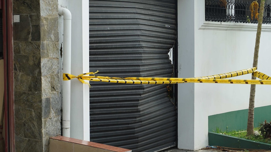 A black garage door is badly dented with yellow crime scene tape in front.