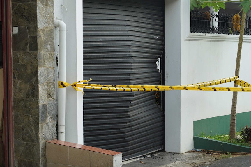 A black garage door is badly dented with yellow crime scene tape in front.