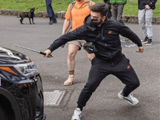 A man wearing black track pants and jacket holds a metal stick in his hand and is about to strike a car.