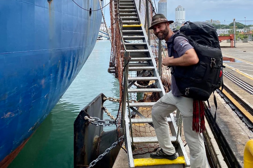 A man with a large backpack stands at the bottom of a staircase onto a cargo ship