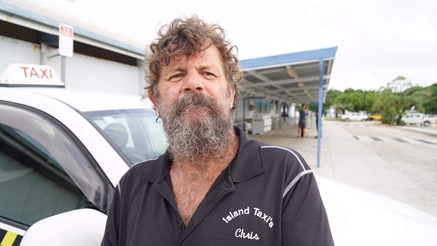 A man with a bushy beard and curly hair standing in front of a taxi at an airport.