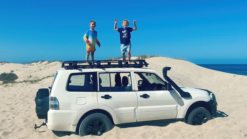 Two young boys stand on top of a 4WD on the sand.
