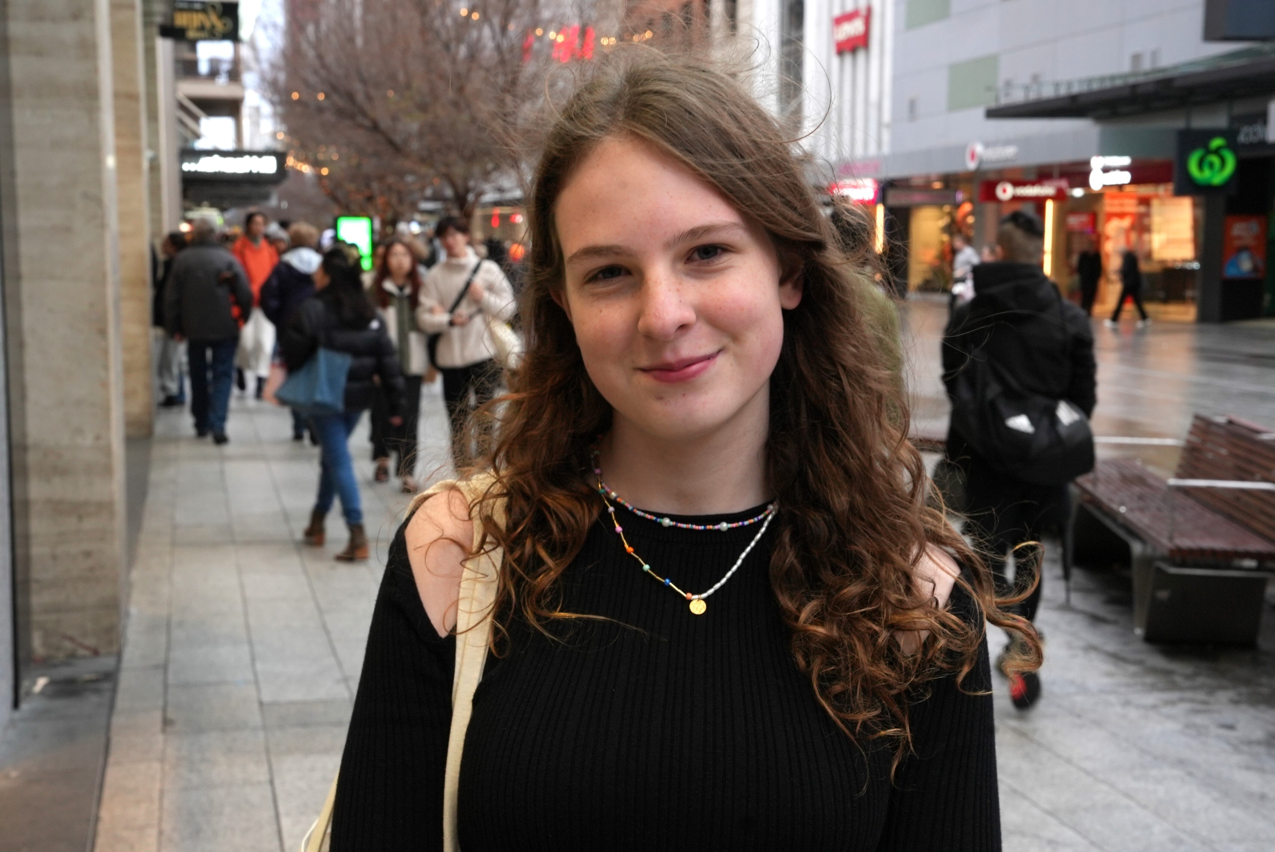 A smiling teenage girl stands in an outdoor shopping mall where it has recently been raining.