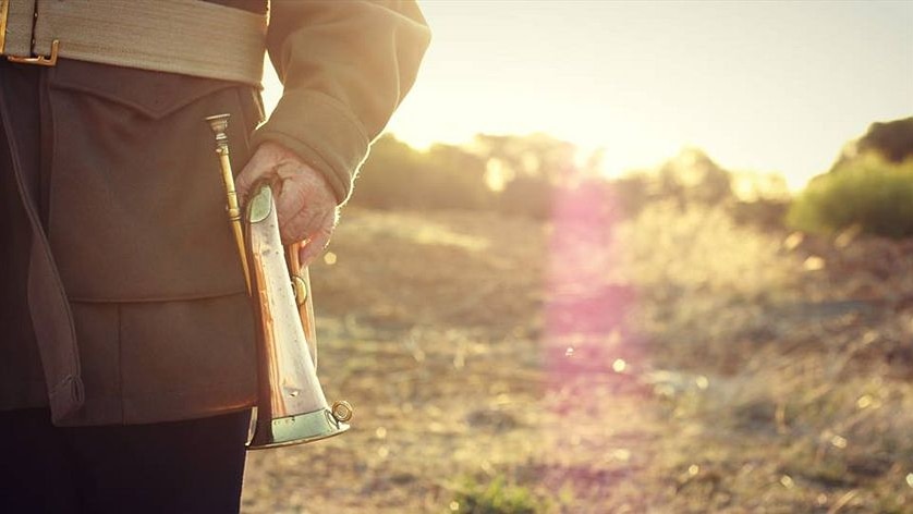 Photo of man dressed in khaki holding a bugle with the sun setting behind him