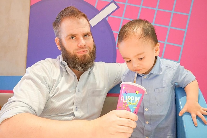 A man with beard smiling to the camera while holding a cup next to a little child.