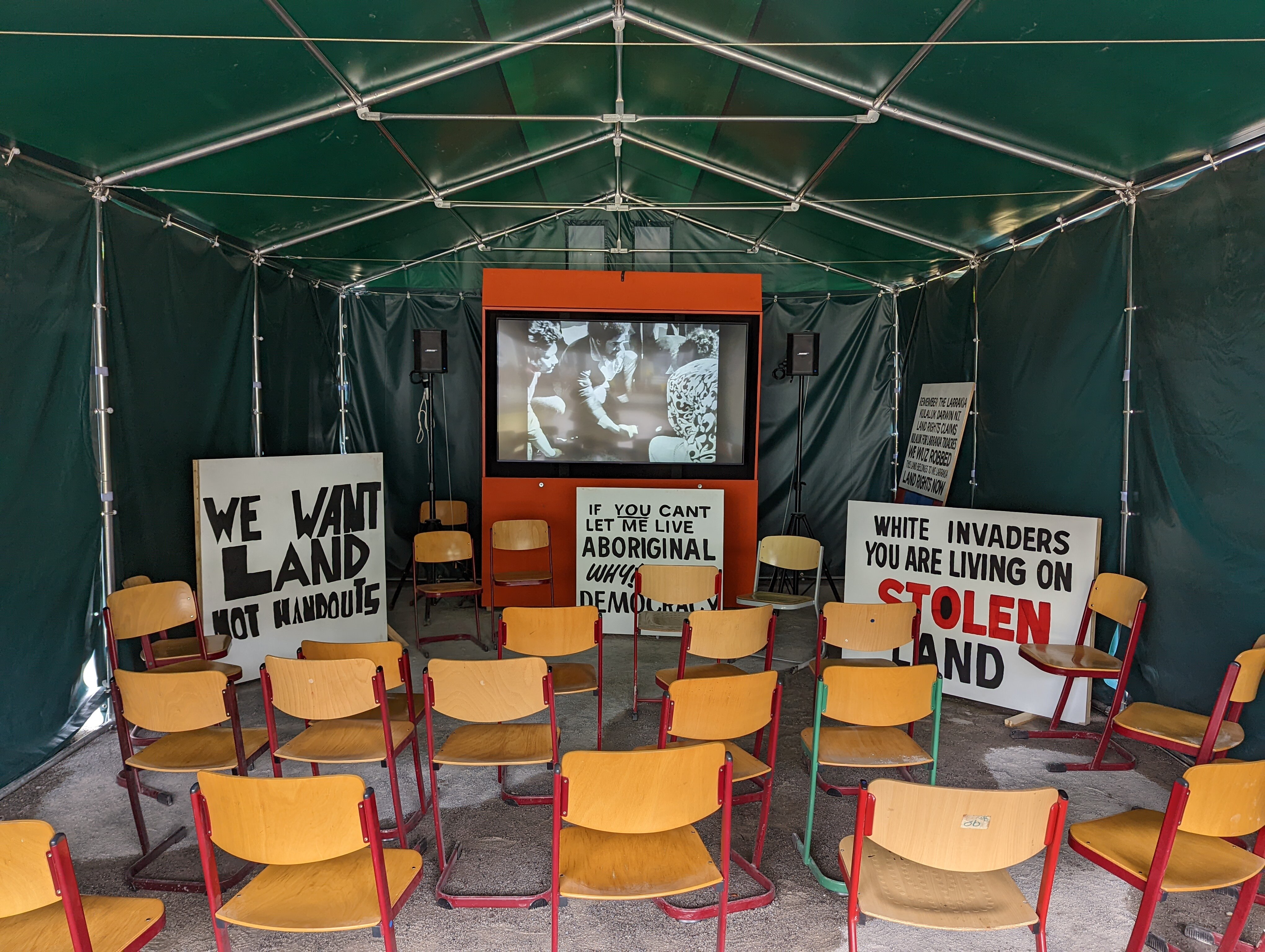 The inside of a large green tent with chairs set in front of a screen featuring black and white video and three protest placards