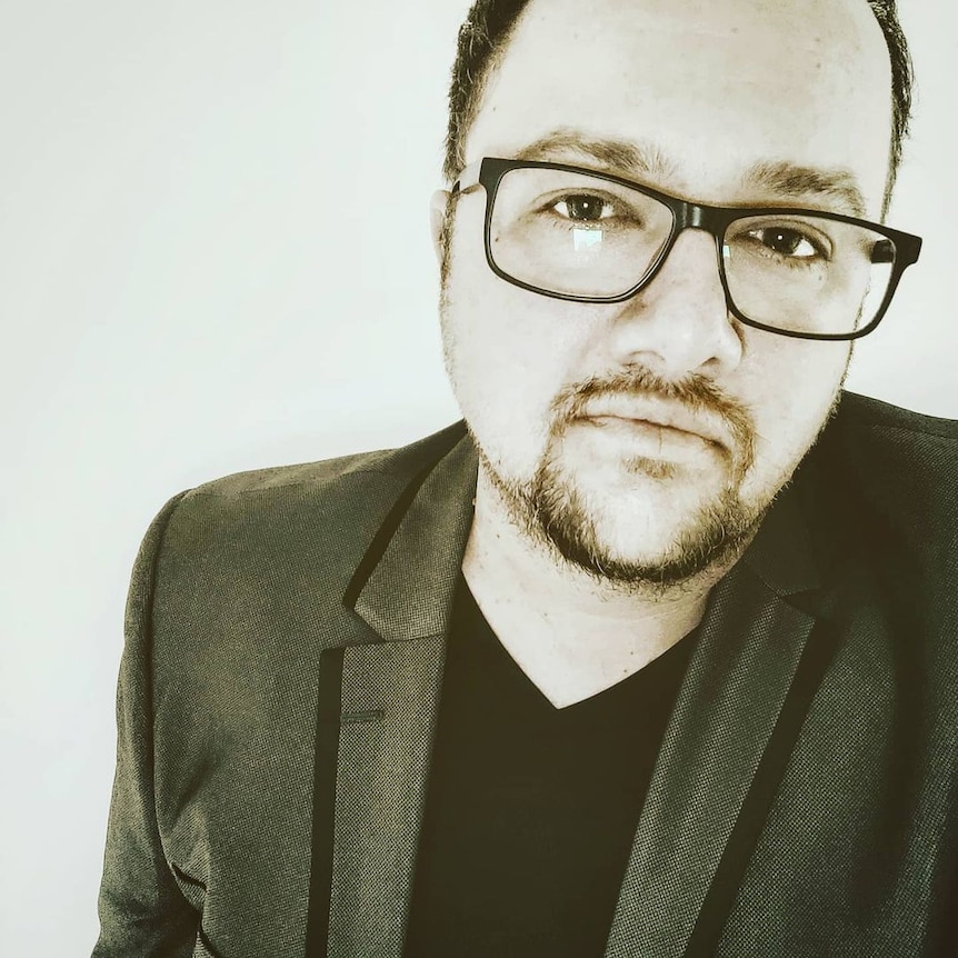 A man with a goatee, prescription glasses, wearing a v neck t-shirt and a blazer in a black-and-white selfie. 
