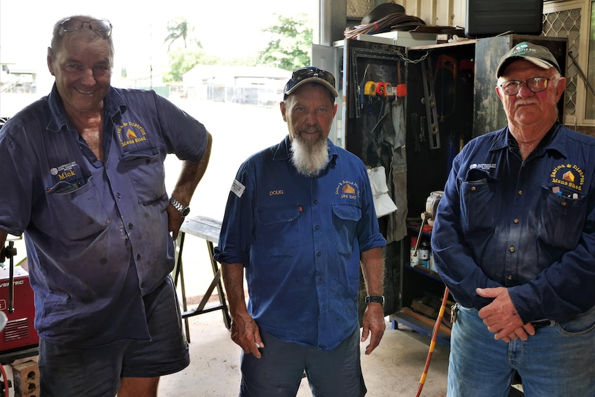 Three men stand together in a shed with the same blue collared work shirts. 