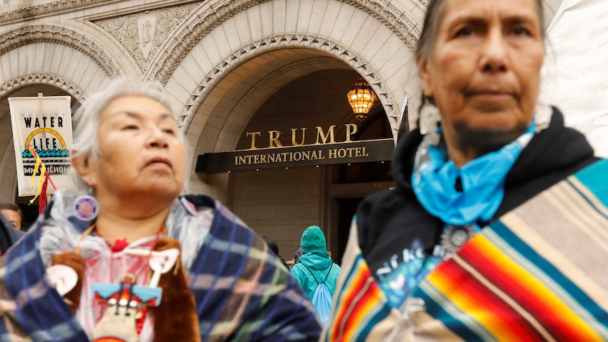 Indigenous women stand, out of focus, in front of the Trump International Hotel.