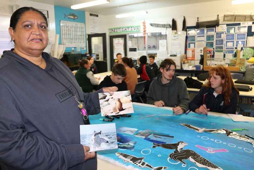 Indigenous woman in foreground with pictures in hand, blue canvas, sea animal pictures, students in background paintingde