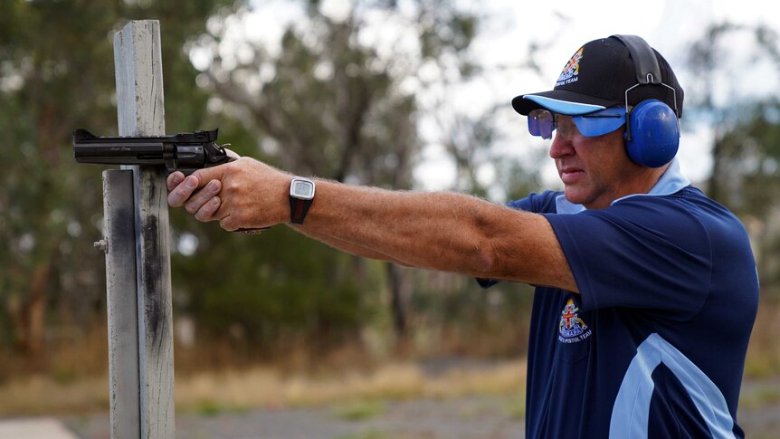 A man wearing a cap and protective earmuffs and glasses points a pistol out in front