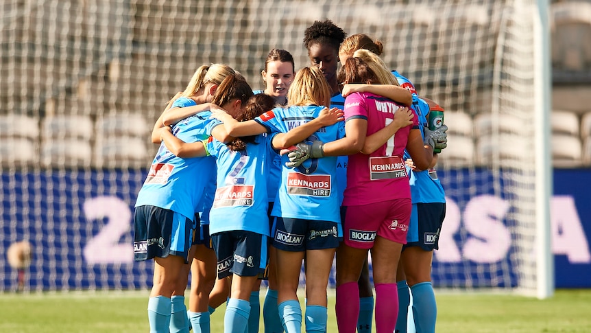Women soccer players wearing light blue jerseys stand in a circle with arms around each other