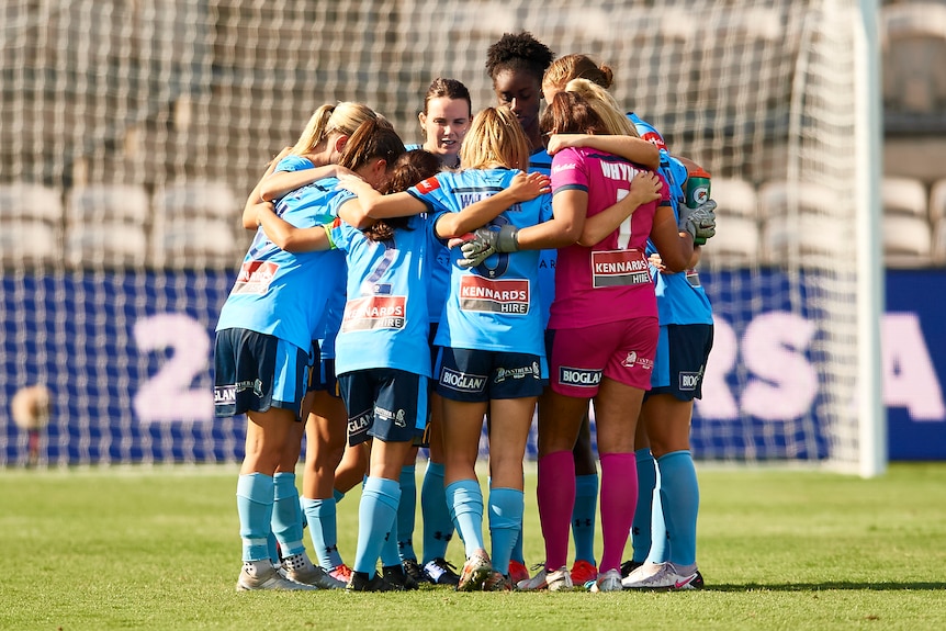 Women soccer players wearing light blue jerseys stand in a circle with arms around each other