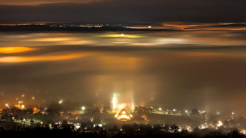 The Australian War Memorial and the lights of the Parliament House flagpole appear through dense fog blanketing Canberra.