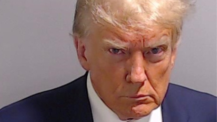 Donald Trump frowns in a mugshot, wearing a suit and red tie