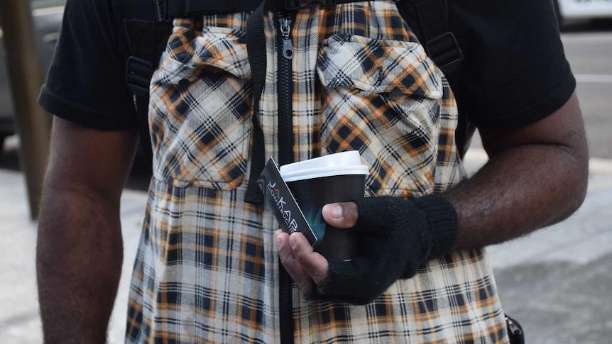 Close up of man's torso, wearing chequered vest, and holding coffee cup and business card in his left hand.