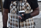 Close up of man's torso, wearing chequered vest, and holding coffee cup and business card in his left hand.