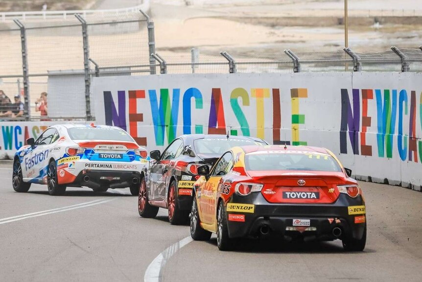 Cars race down a street lined with fencing and colourful Newcastle signs, above Newcastle beach.