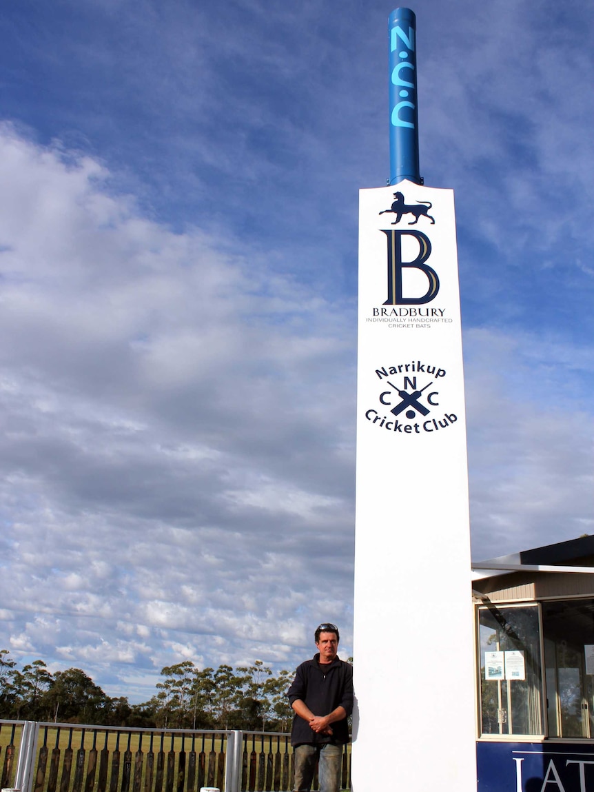 A man standing next to a giant cricket bat in front of an oval