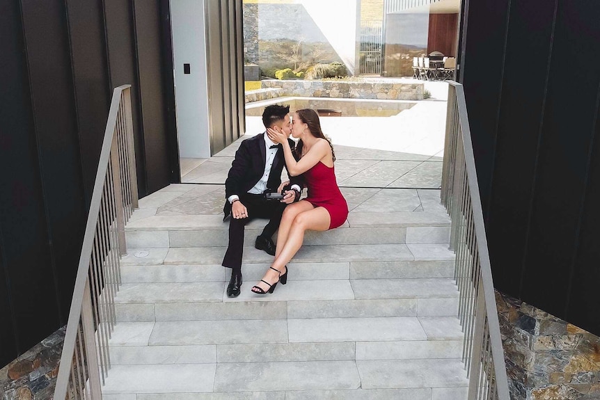 Brandon Jackson and Lara Markham sit in formal attire, kissing on the steps of a luxury house in Gerringong, shot from a drone.