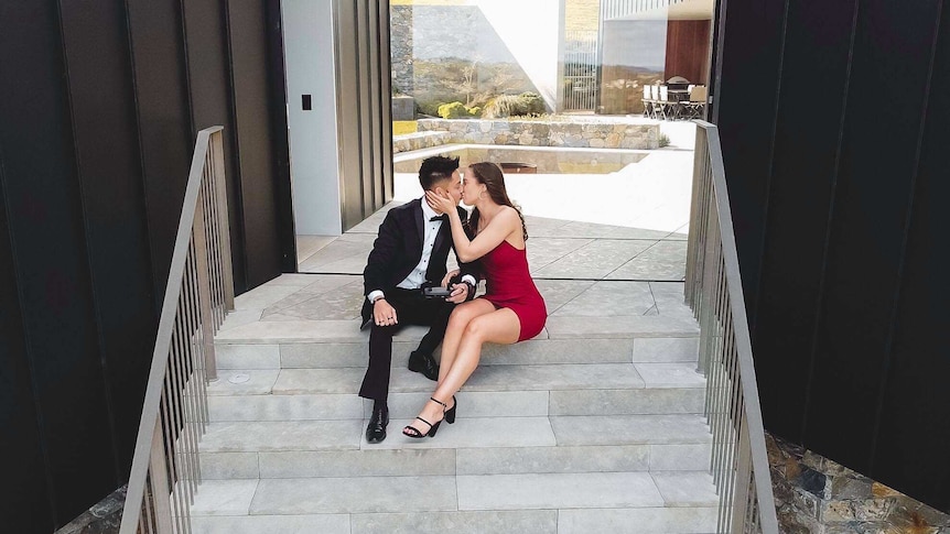 Brandon Jackson and Lara Markham sit in formal attire, kissing on the steps of a luxury house in Gerringong, shot from a drone.
