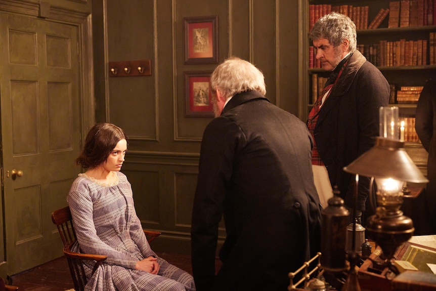 A young white woman with dark hair dressed in 1800s attire sits being reprimanded in a chair by two old grey-haired men.