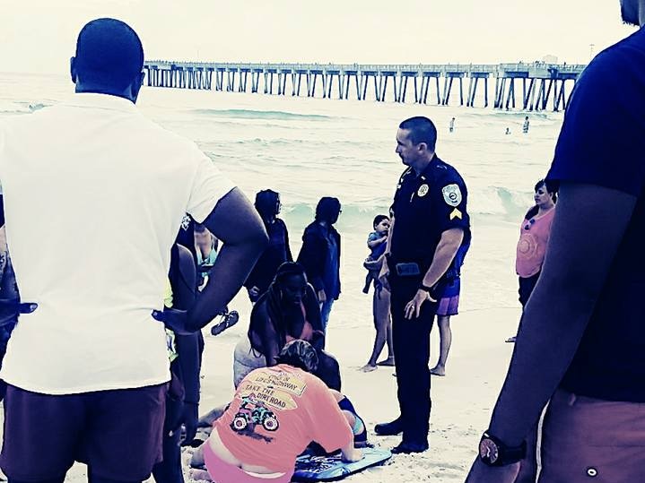 People, including a police officer in uniform, gather around a group of people on the beach assisting someone