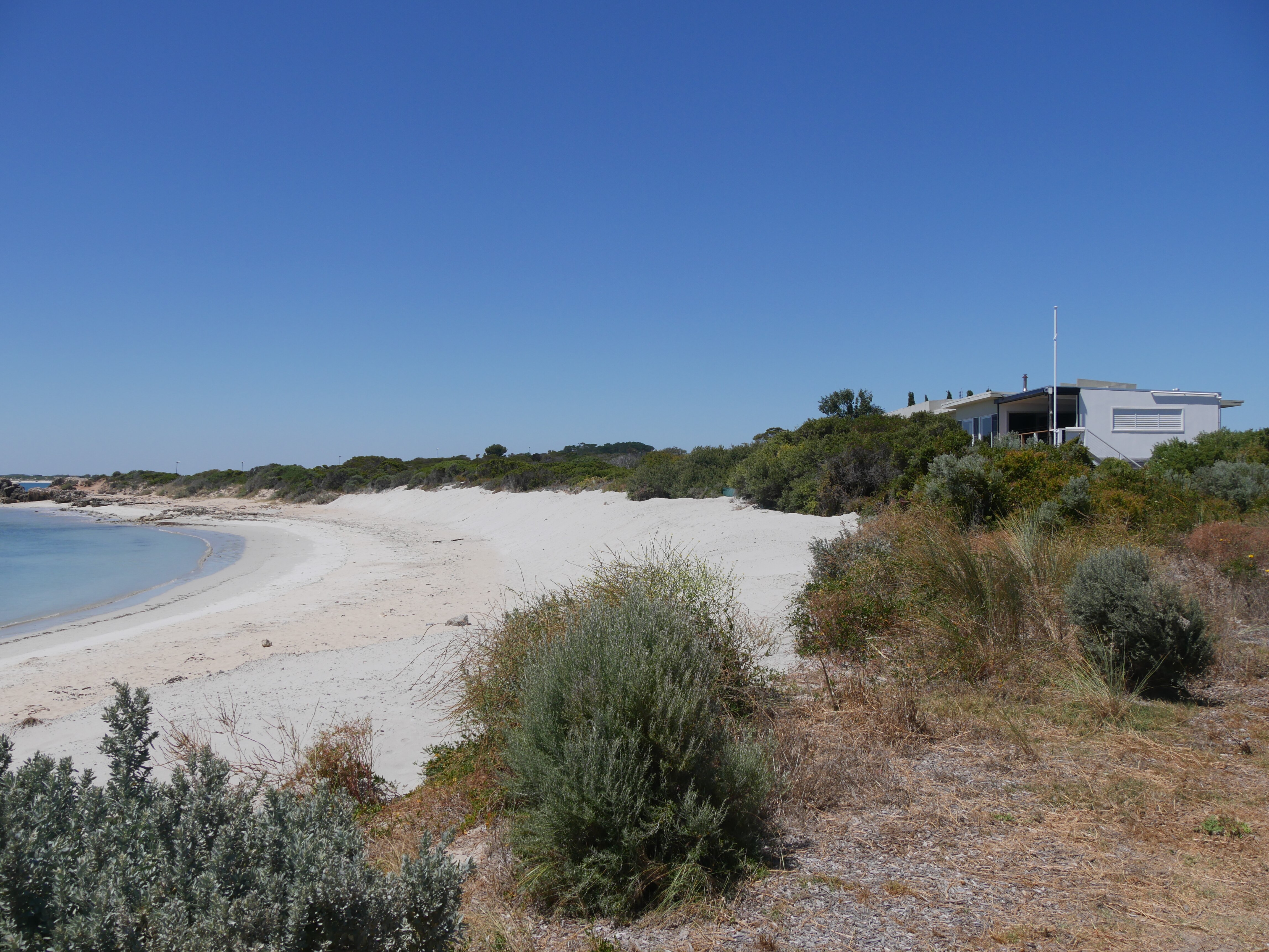 A beach with water to the left of the shot, a sand dune in the centre, and houses atop the dune