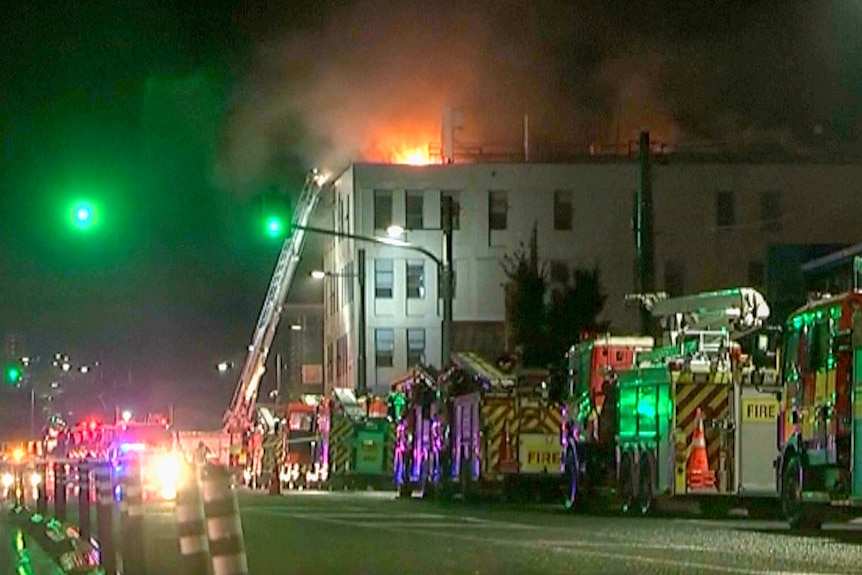 Flames rising out of a building with several firetrucks lined outside. 