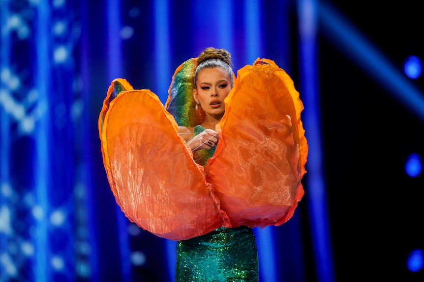 Rikkie Kolle wearing a tick green glittery dress with orange tulip petals extending from her hips 