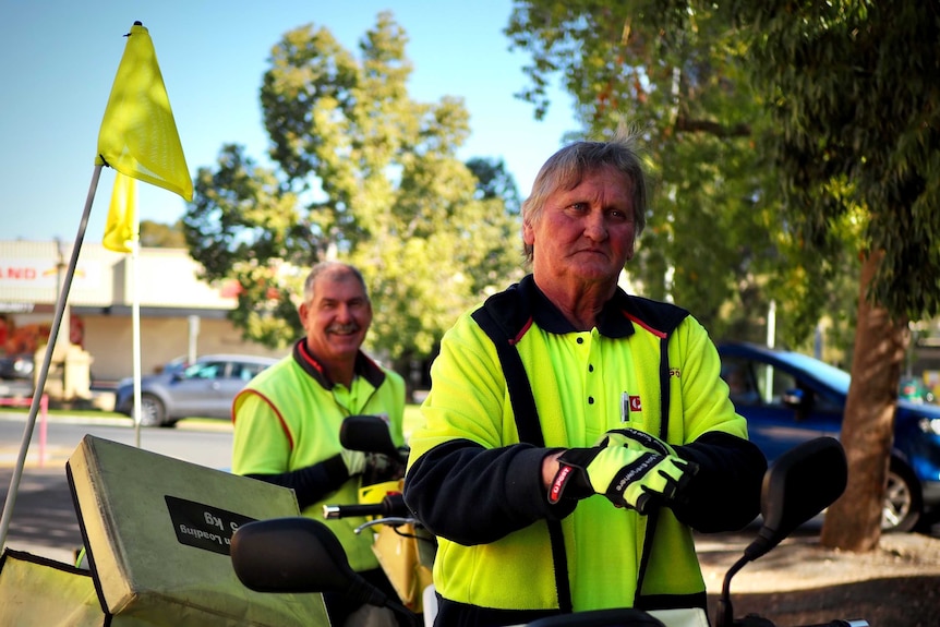 Two postmen wearing fluro vest put on their gloves outside, standing near their motorbikes