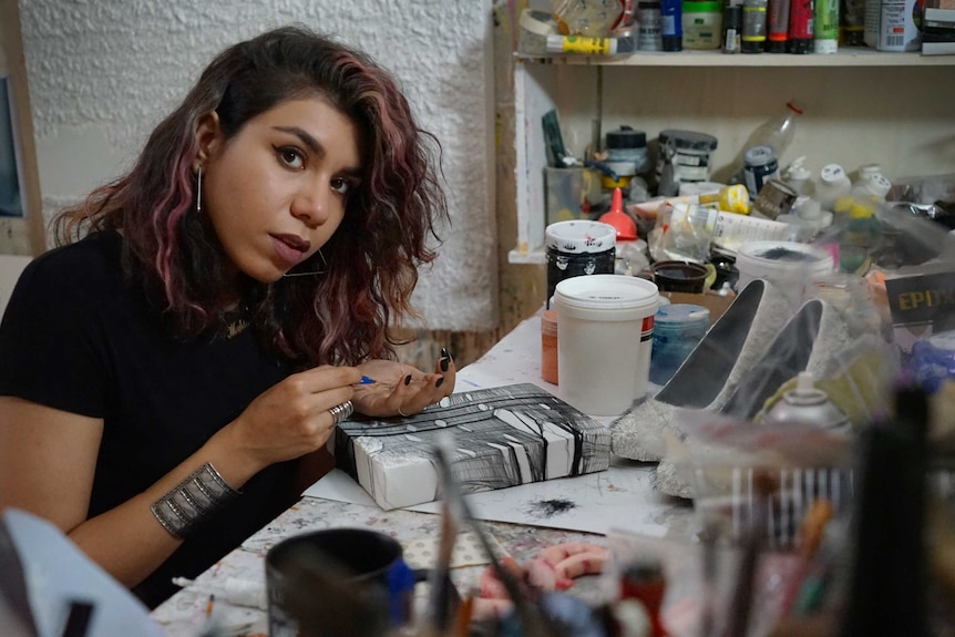 A woman sits at a table with craft supplies in front of her and looks at the camera.