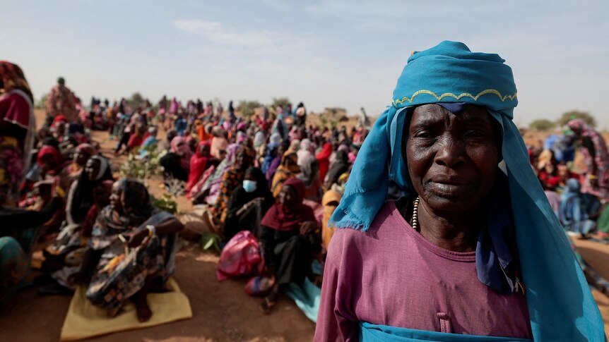 A Sudanese refugee stands in front of rows of people sitting on the ground in Chad.