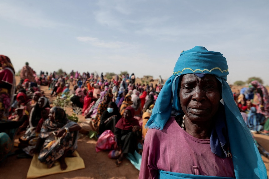 A Sudanese refugee stands in front of rows of people sitting on the ground in Chad.
