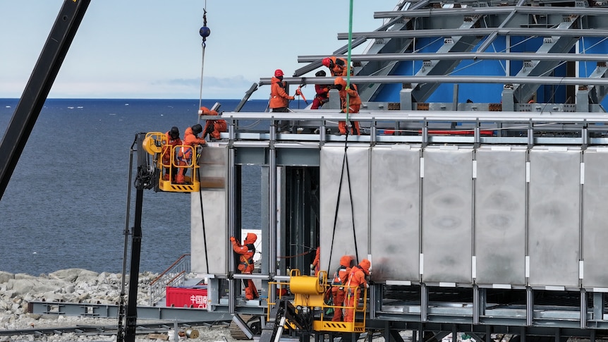 workers in orange protective gear construct a steel building frame overlooking the sea in Antarctica.