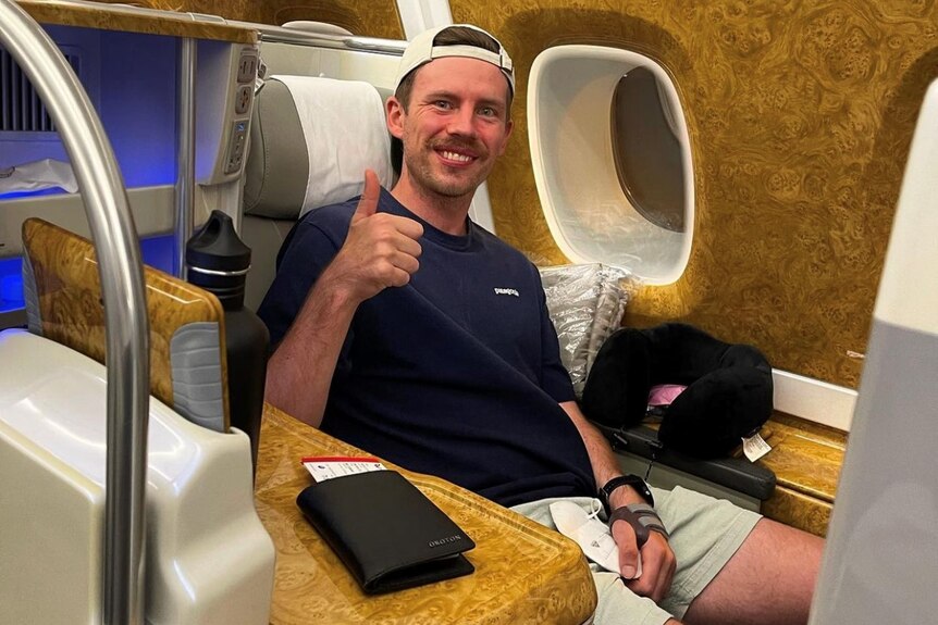 Chris Ross sits in a business class airplane seat and gives the thumbs up