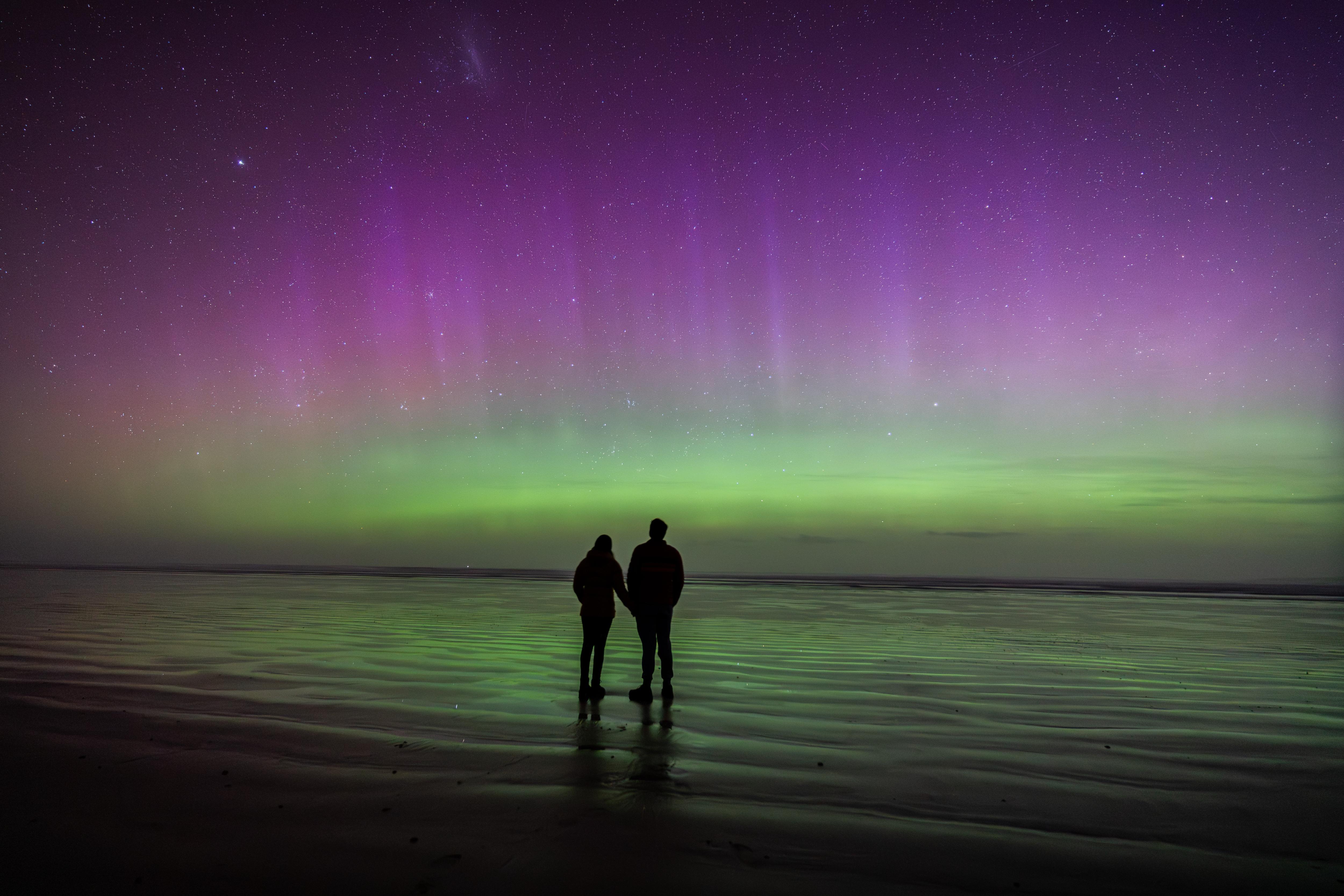 Um céu roxo e verde com duas silhuetas na praia.