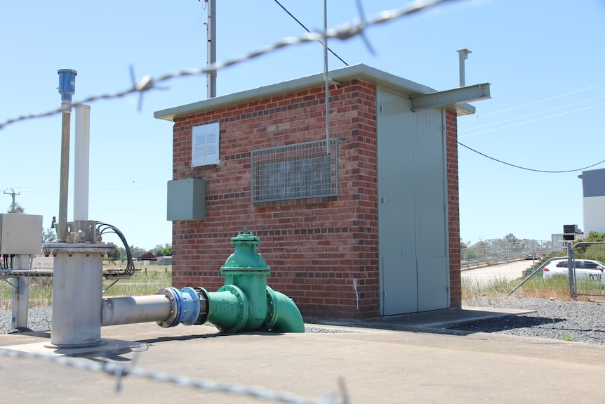 A square brick building and a large green pump, both surrounded by a barbed wire fence.