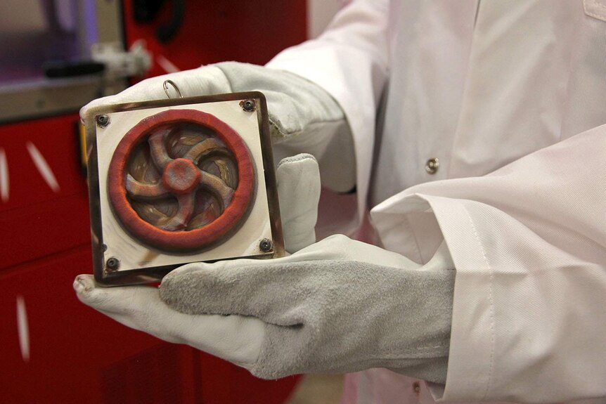 A person in a lab coat holds a copper flywheel that has just been printed by the SPEE3D printer.