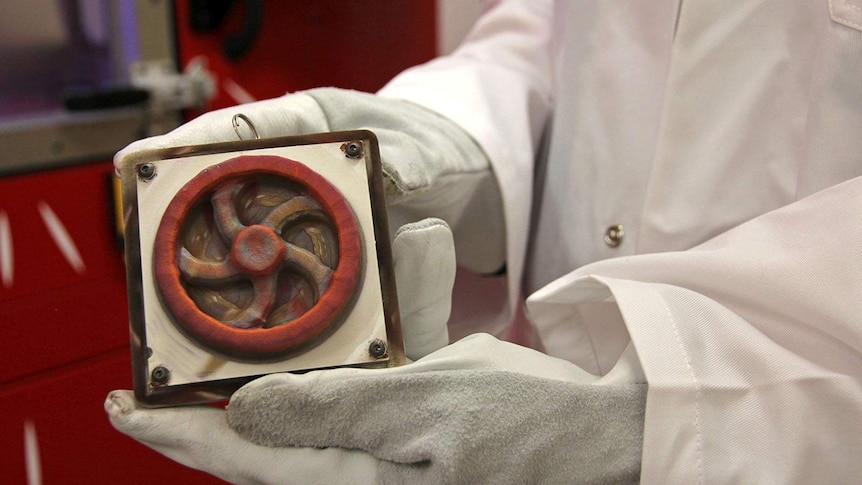 A person in a lab coat holds a copper flywheel that has just been printed by the SPEE3D printer.