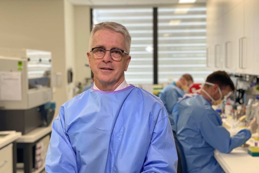 A man with white hair and wearing glasses and blue scrubs smiles in a labratory.