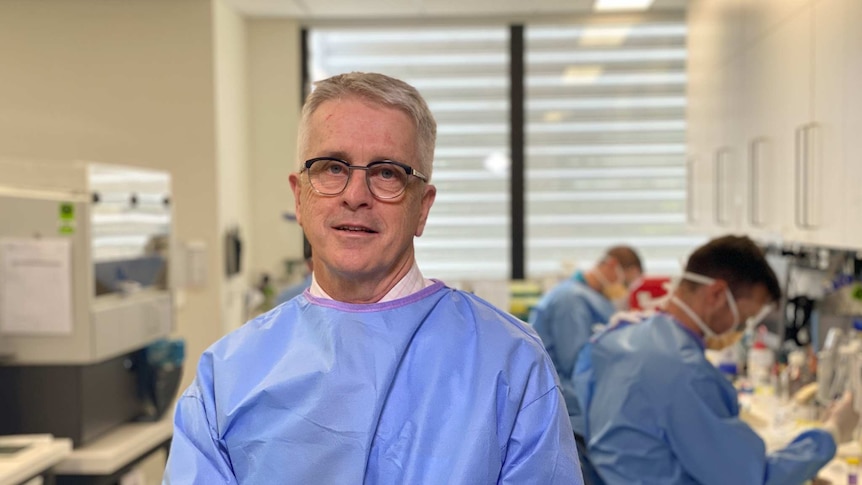 A man with white hair and wearing glasses and blue scrubs smiles in a labratory.