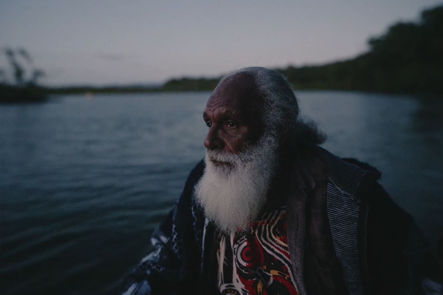 An older Aboriginal man with a grey bear in a boat at sunrise against the backdrop of water.