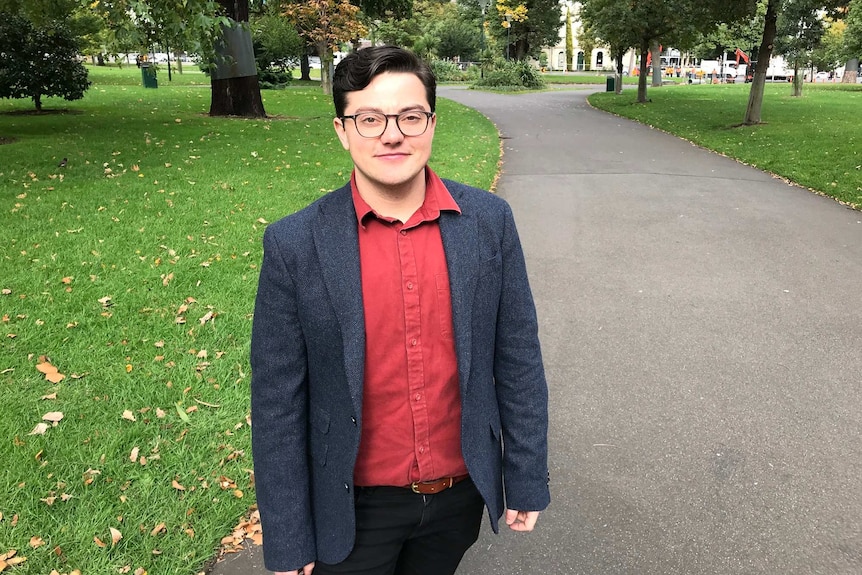 Sam Elkin stands in a park wearing glasses, a red shirt and grey blazer.