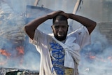 A man looks distressed, with his hands on his head, as he stands in front of a burning building.