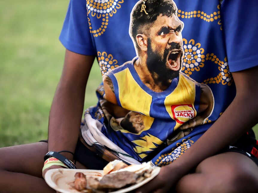 Bright coloured t-shirt of footballer Liam Ryan worn by young Aborigina girl holding a plate of food