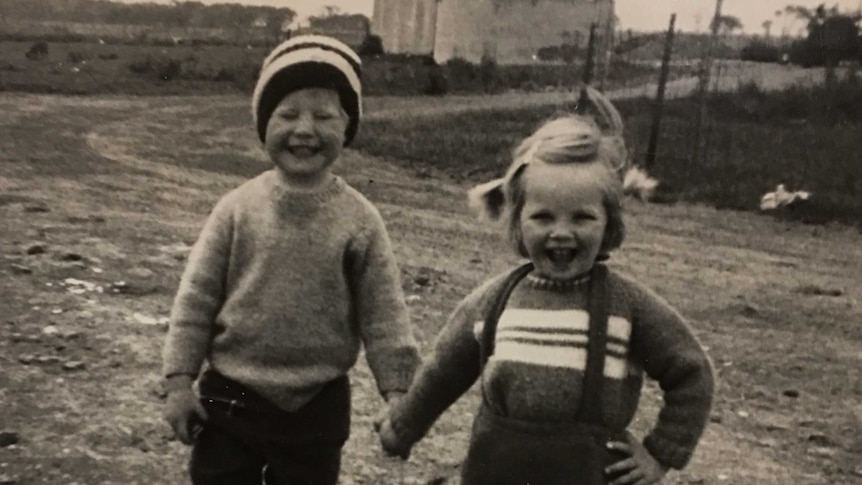 Black and white grainy photograph depicting two grinning children holding hands in a farmyard setting